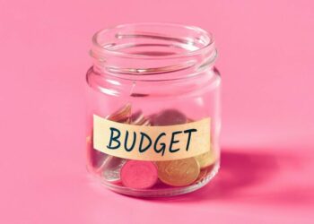 A clear glass jar labeled "BUDGET" filled with coins, set against a solid pink background.